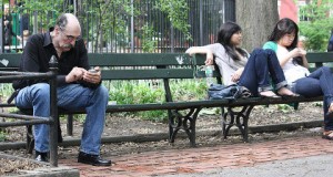 older man sitting on a park bench texting two young women sit at the opposite end of the bench