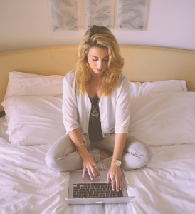young woman sitting on her bed typing on a laptop